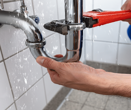 Plombier réparant une fuite d'eau sur un siphon chromé sous un lavabo avec une clé serre-tube rouge.