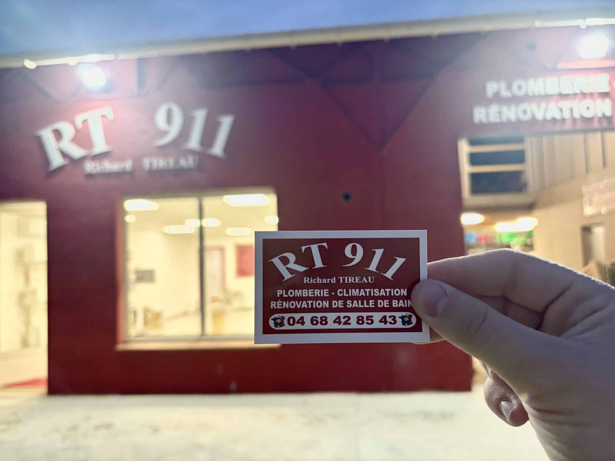 Main tenant une carte de visite RT 911 Richard TIREAU Plomberie Climatisation 04 68 42 85 43 devant un bâtiment rouge
